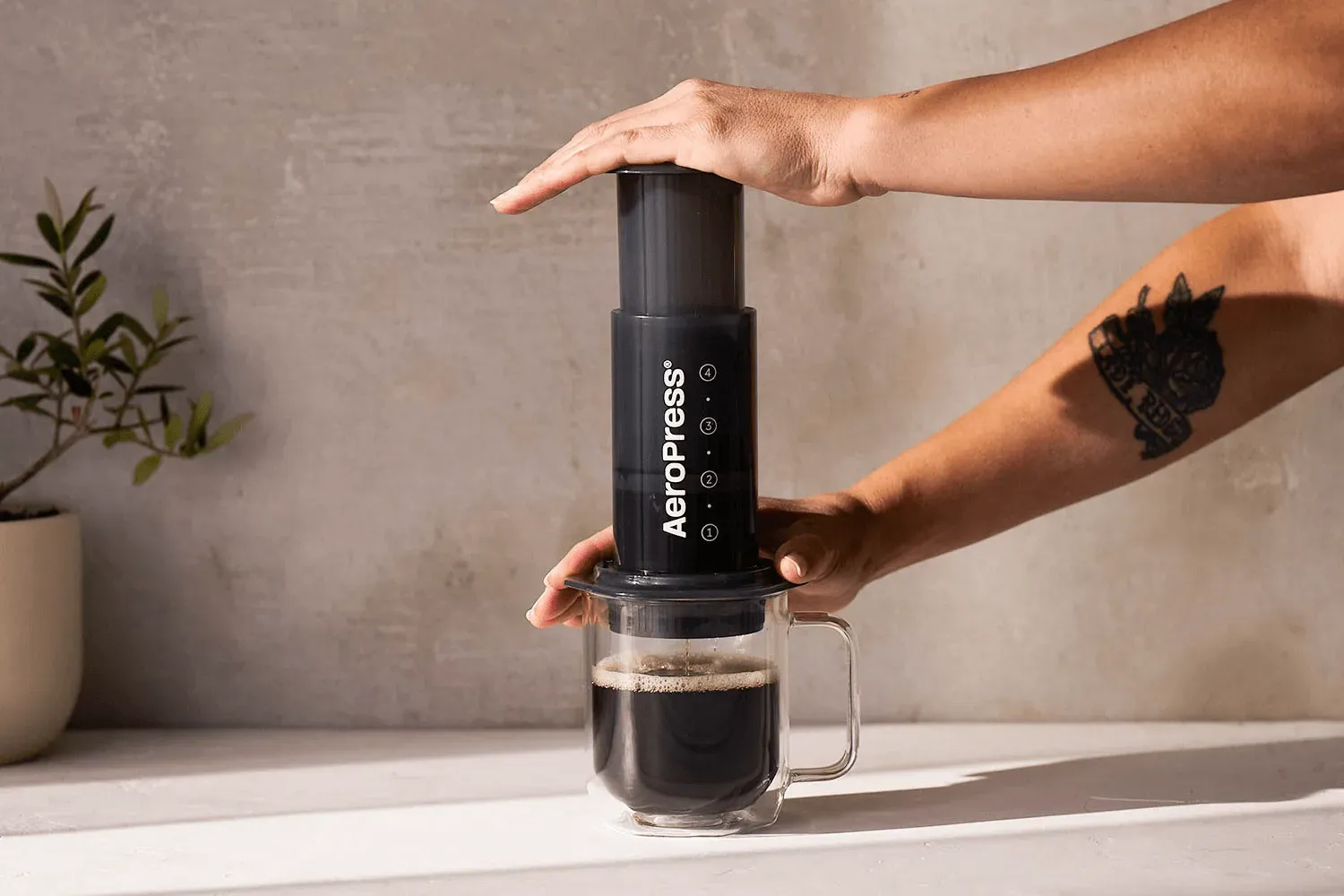 Des mains enfoncent le piston d'une AeroPress Original dans un mug en verre rempli de café, sur un plan de travail au soleil