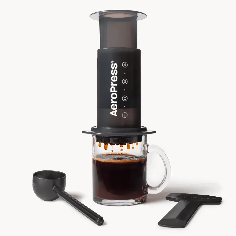 AeroPress Original coffee maker pressed onto a glass mug, with its scoop and stirrer beside it