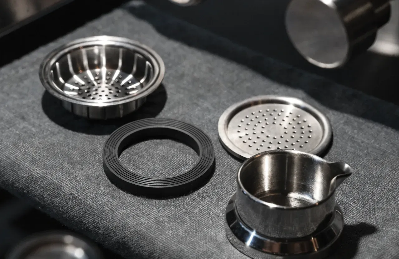 Un porte-filtre, un joint de groupe de rechange, une douchette et deux paniers filtres posés à côté d'une machine espresso