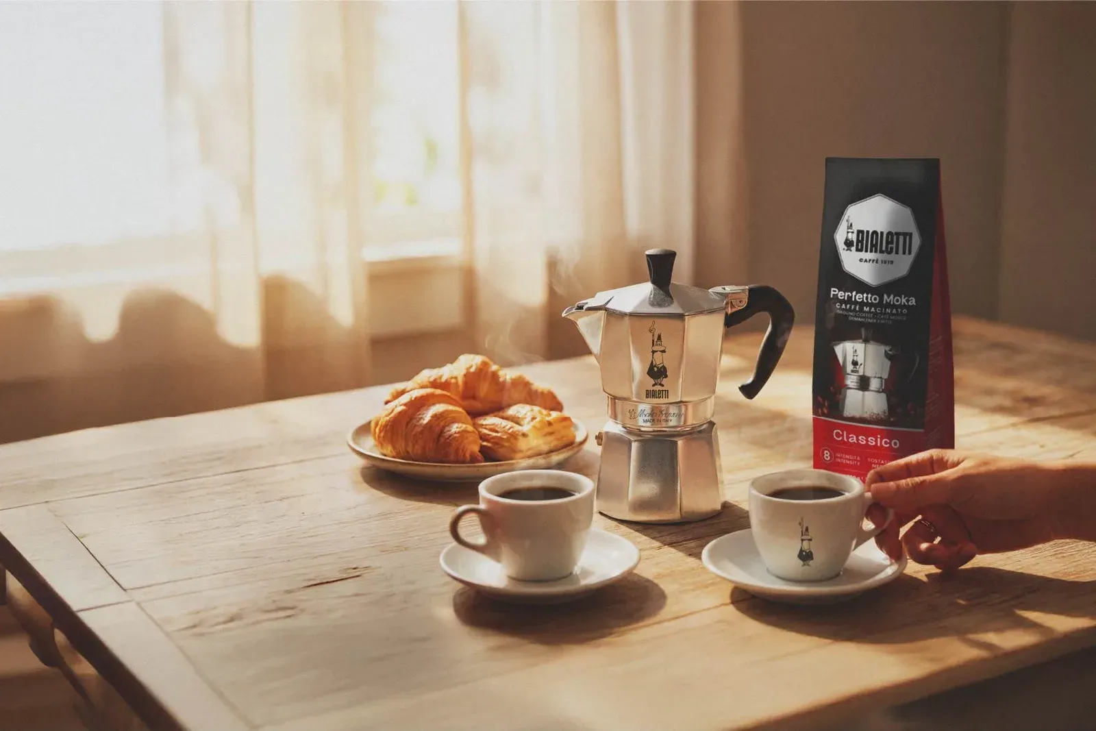 Une Bialetti Moka Express sur une table de petit déjeuner en bois, avec deux tasses de café et une assiette de croissants, dans la lumière du matin