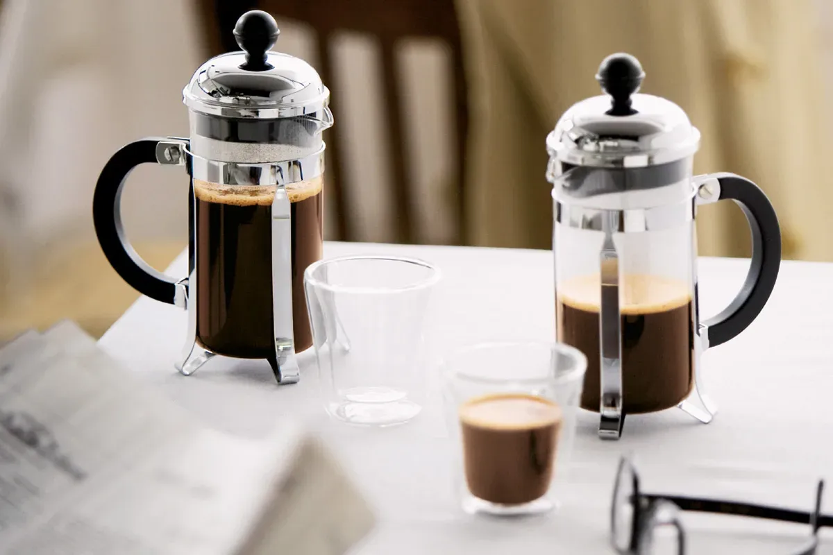 Deux cafetières à piston Chambord chromées remplies de café sur une nappe blanche, avec de petits verres, un journal et des lunettes