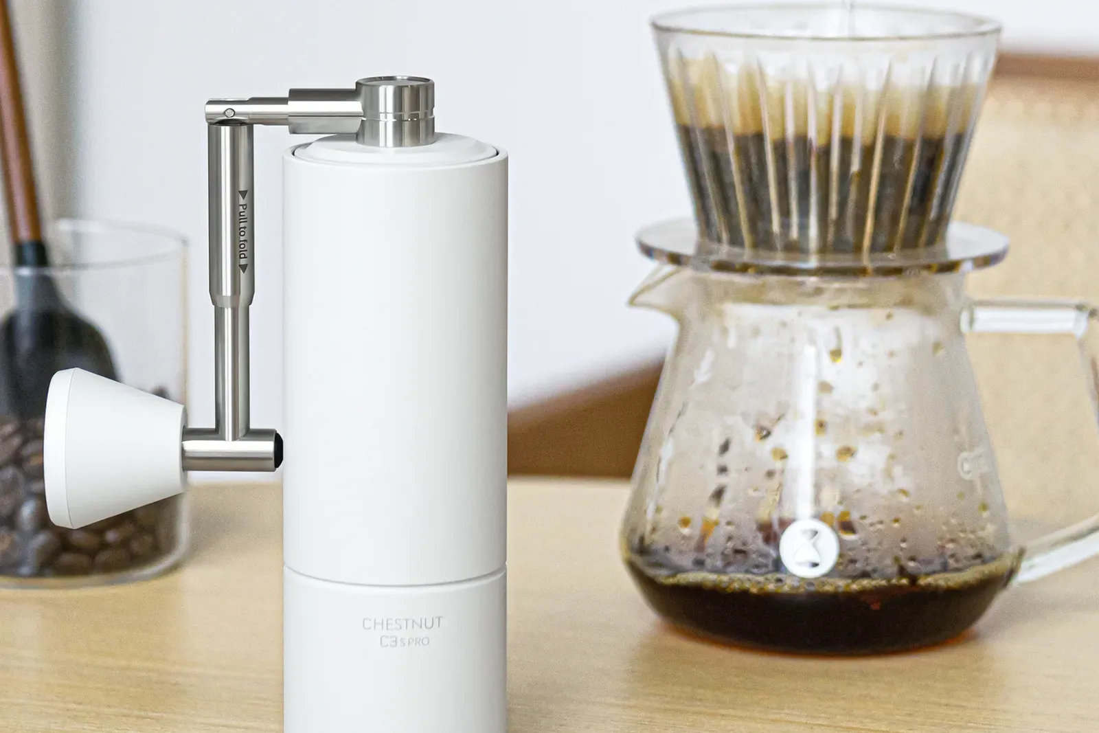 Un moulin manuel Timemore Chestnut C3S Pro blanc à côté d'un dripper et d'une carafe en verre remplie de café