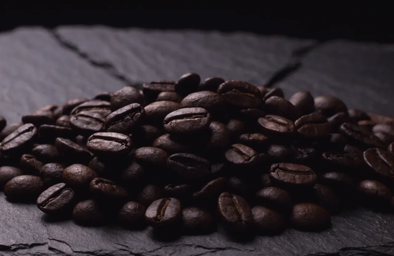 Des grains de café en torréfaction foncée au léger voile d'huile, sortant d'un sachet en papier sur un plan de travail sombre