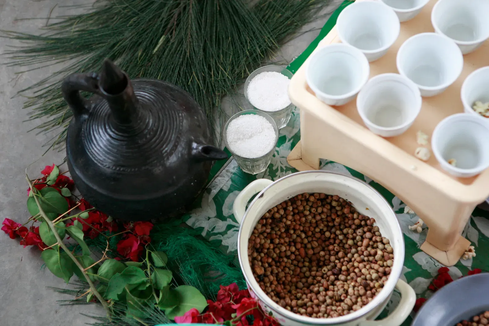 Une jebena d'argile noire posée sur de l'herbe et des bougainvilliers, à côté d'un bol de grains torréfiés et d'un plateau de petites tasses blanches