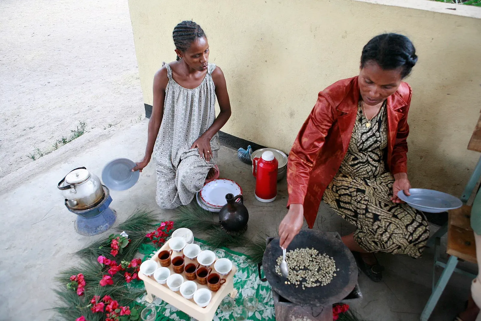 Une femme torréfie du café vert dans une poêle plate sur des braises, des petites tasses et une jebena d'argile posées sur de l'herbe fraîche à côté d'elle