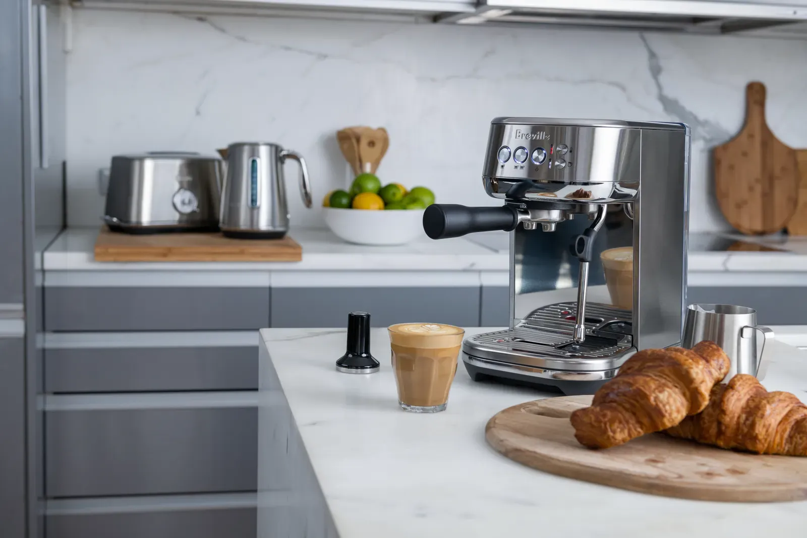Une machine espresso Breville Bambino Plus sur un plan de travail en marbre, à côté d'un verre de café au lait et d'une planche de croissants