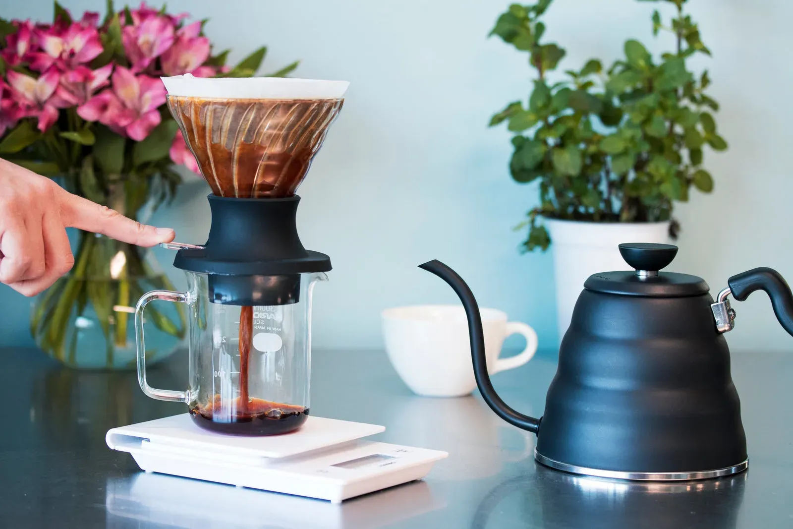 Un doigt appuie sur le levier d'un dripper Hario Switch en verre, et le café s'écoule dans une verseuse posée sur une balance