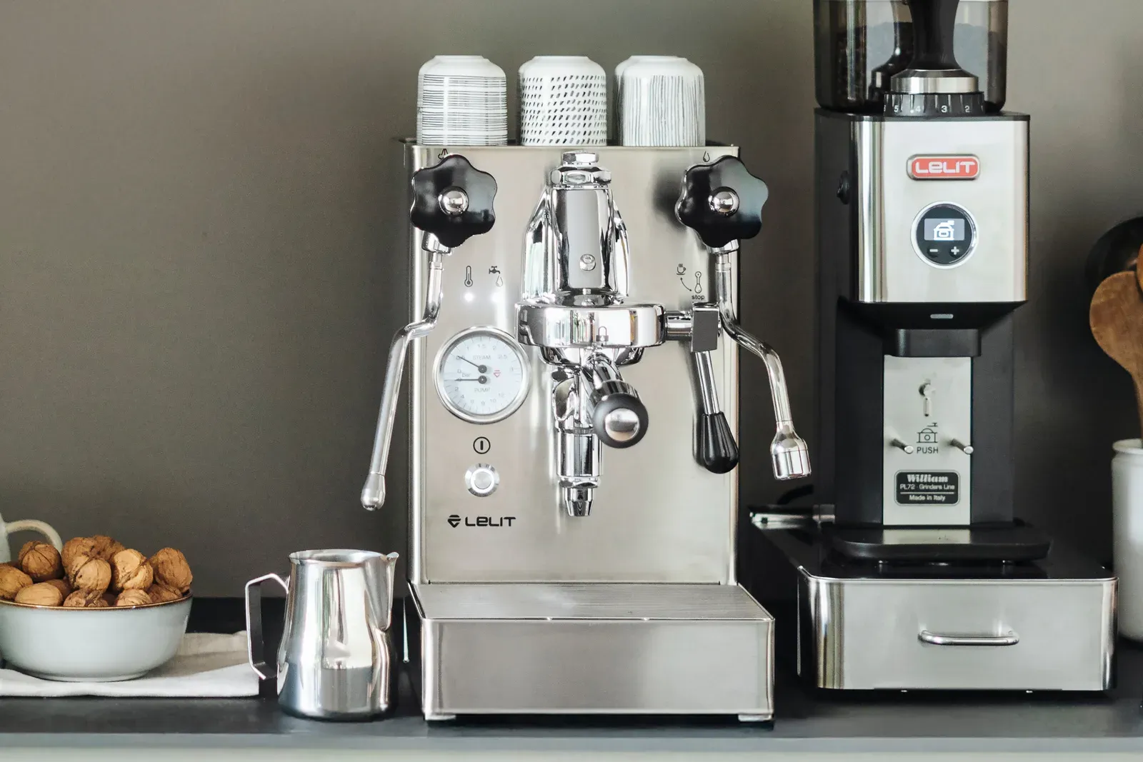 Une machine espresso Lelit Mara X en inox sur un plan de travail, entre un pot à lait et un moulin Lelit, des tasses posées dessus