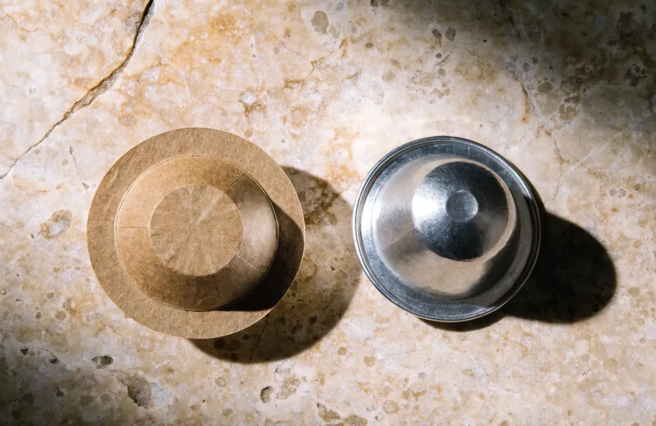 Une dosette ESE en papier et une capsule en aluminium côte à côte sur un plan de travail, avec un panier à dosette posé dans un porte-filtre derrière