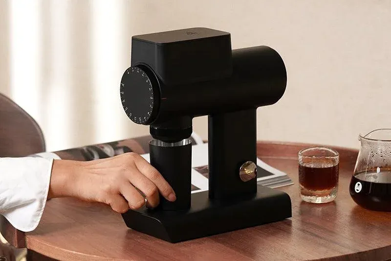 Une main place le réceptacle sous un moulin Timemore Sculptor 064S noir, sur une table en bois à côté d'un verre de café filtre
